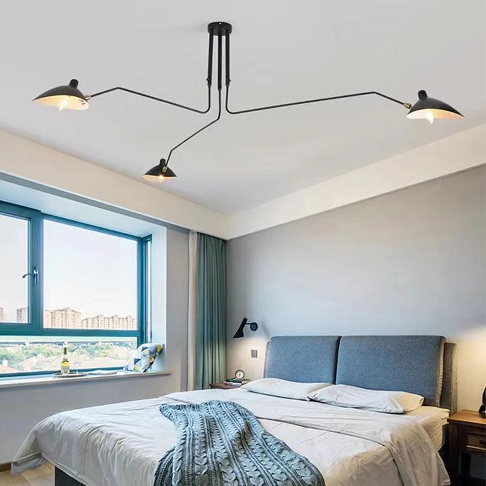 A modern bedroom showcases a grey bed with a blue blanket, accompanied by a laptop. The room is crowned by a Sleek Industrial Pendant Light with elegant brass accents. A large window on the left offers city views and a wall light above the bedside table adds warmth.