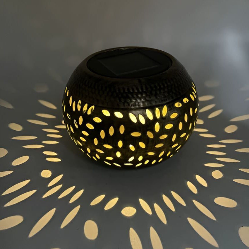 The Solar Orb Lamp features a round, hollow iron design with intricate cut-out patterns that emit a warm yellow light, casting decorative shapes onto nearby surfaces. Ideal as a solar patio light, it adds both beauty and ambiance to your outdoor space.