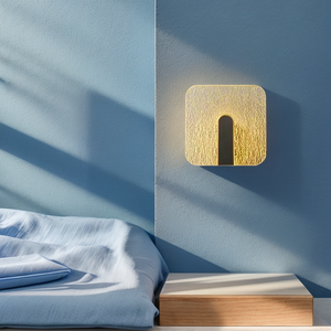 The Beautiful Wall Sconce, featuring a textured and illuminated surface, is mounted above a minimalistic bed and bedside table set against a blue wall.