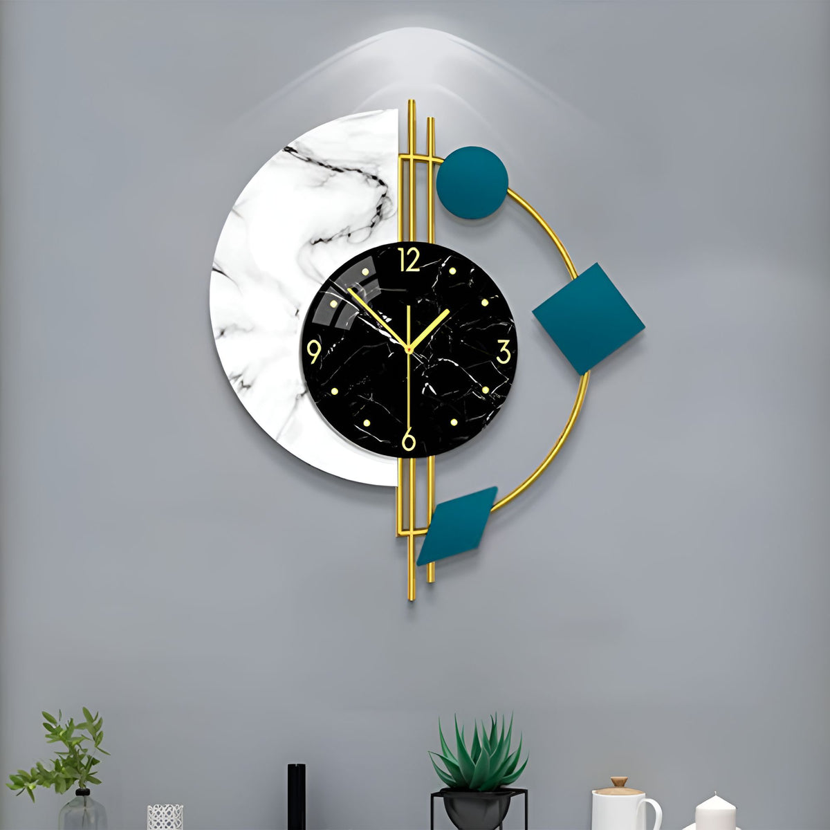 The Modernist Constellation Accent Clock features a black marble face, gold accents, and geometric teal and white shapes—an eye-catching statement piece for any space, shown mounted on a grey wall above a desk with plants and decor.