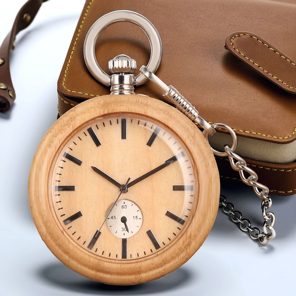 The Wood Pocket Watch, featuring black hour markers and a small seconds dial on solid wood, is attached to a chain and sits next to its brown leather case.