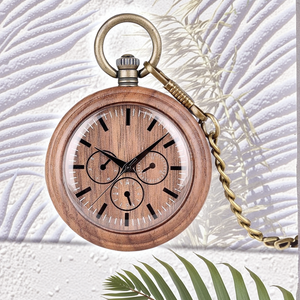 The Handmade Wood Pocket Watch with a brass chain rests on a textured white surface, surrounded by dancing palm leaf shadows and a green palm frond in the foreground.