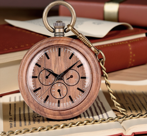 The Handmade Wood Pocket Watch in natural walnut, with its elegant chain, rests atop an open book, surrounded by closed books and scattered papers.