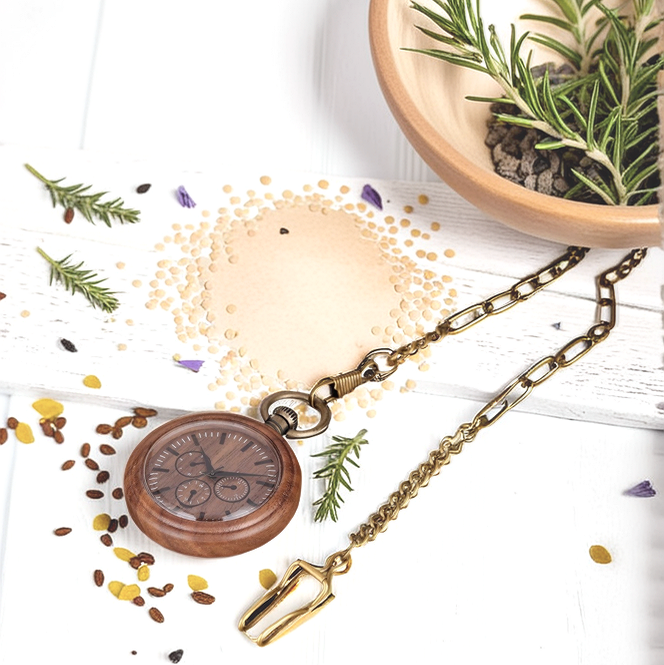 A Handmade Wood Pocket Watch with a brass chain rests on a white surface next to a bowl of herbs, scattered seeds, rosemary sprigs, and small decorative accents.