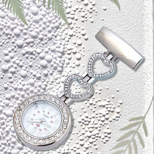 The Luxe Fob Pocket Watch, featuring a silver finish with a heart pattern and rhinestone accents, is displayed on a textured white background amid green fern leaves.