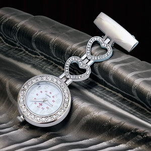 The Luxe Fob Pocket Watch is a silver nurse fob watch with a round face, crystal accents, and three heart-shaped links featuring a delicate heart pattern, displayed on textured silver and black fabric.