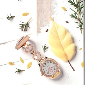 The Luxe Fob Pocket Watch with gold heart-shaped links and crystal accents rests on a white wooden surface amid yellow petals, green sprigs, and a delicate yellow feather.