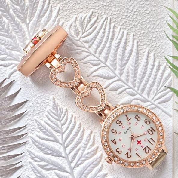The Luxe Fob Pocket Watch, a gold nurse fob watch with a jeweled bezel, crystal accents, and two heart-shaped links, rests on a white textured leaf background with green leaf accents.
