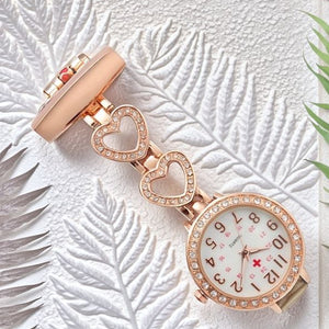 The Luxe Fob Pocket Watch, a gold nurse fob watch with a jeweled bezel, crystal accents, and two heart-shaped links, rests on a white textured leaf background with green leaf accents.