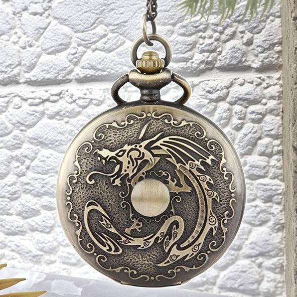 The Dragon Pocket Watch is a round, antique brass timepiece featuring an ornate dragon design engraved on the cover, suspended from a chain against a textured white background.