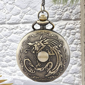 The Dragon Pocket Watch is a round, antique brass timepiece featuring an ornate dragon design engraved on the cover, suspended from a chain against a textured white background.
