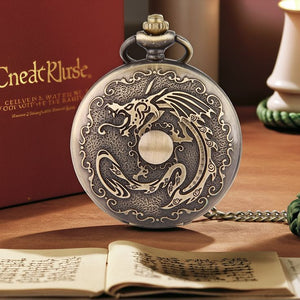 The Dragon Pocket Watch, featuring an ornate dragon design on its antique brass case, rests on a table beside an open book and a red box.