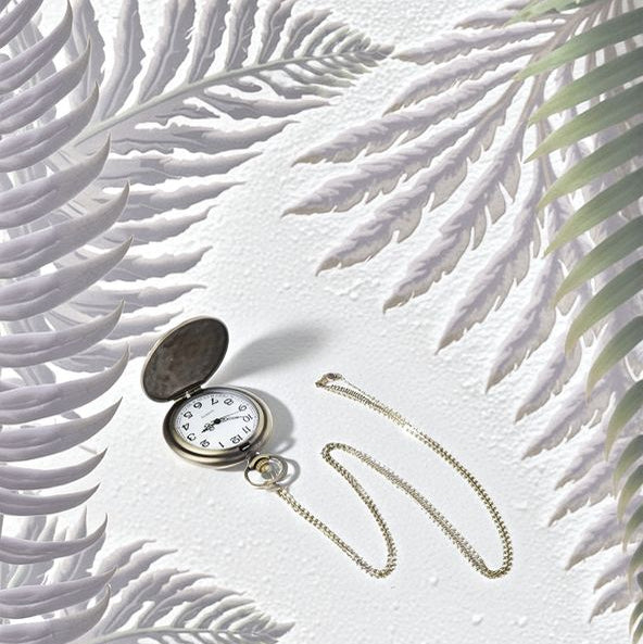 The Dragon Pocket Watch with a gold chain lies open on a textured white surface, surrounded by light purple and green fern leaves.