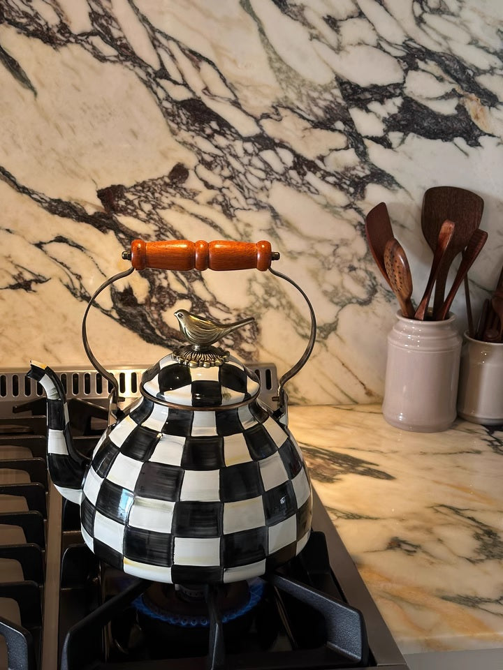 The Vintage Black and White Kettle, featuring a unique checkered pattern, sits on a gas stove with a blue flame beneath it; nearby, utensils are arranged in a ceramic jar on the marble countertop.