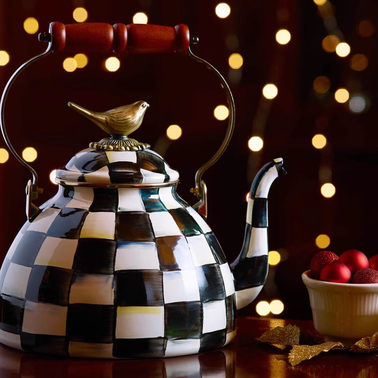 The Vintage Black and White Kettle with a checkered pattern and sparrow-shaped knob sits on a table next to a bowl of red ornaments, as blurred lights glow softly in the background.