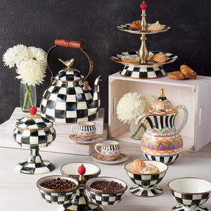 Displayed among treats, white flowers, and wooden crates is the Vintage Black and White Kettle with a sparrow-shaped knob, alongside a black and white checkered tea set of cups and bowls.