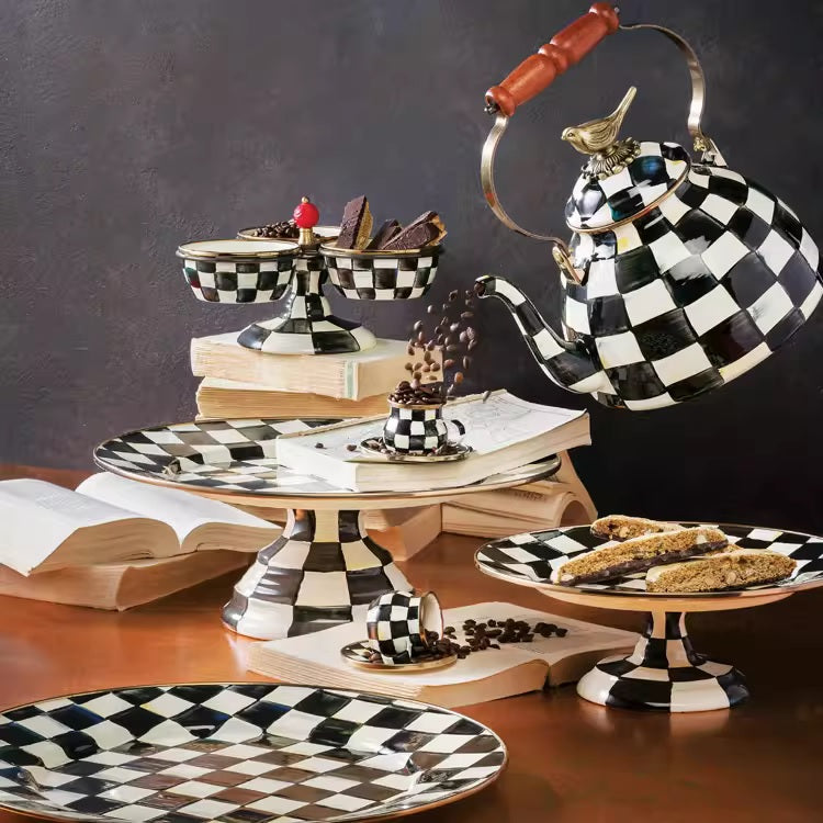 The Vintage Black and White Kettle, featuring a sparrow-shaped knob, pours coffee into a matching cup amid checkered plates and dessert stands with pastries on a table adorned with open books.