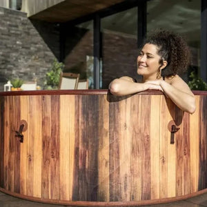 A woman with curly hair smiles as she relaxes in the Outdoor Spa Hot Tub, surrounded by a modern building and lush plants, creating the perfect outdoor spa experience.
