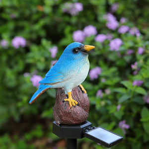 A charming Solar Bird Light featuring a blue and white bird with a yellow beak perched on a stump, set in a garden surrounded by purple flowers and lush green foliage.