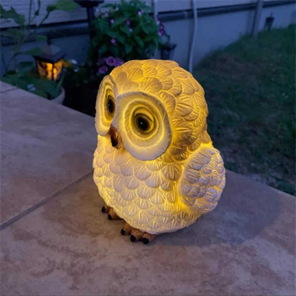 A Solar Owl Light, shaped like a yellow owl, glows on an outdoor table in the evening, serving as a charming garden decoration with plants and a lantern visible in the background.