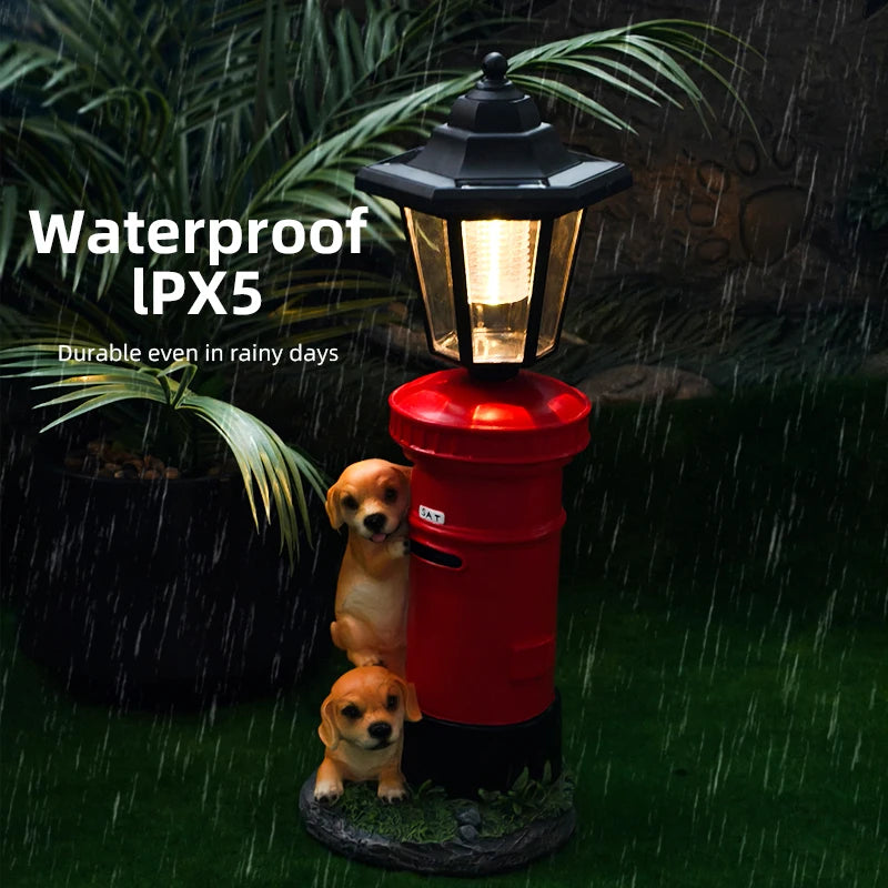 Solar Dog Light designed as a red postbox with two puppy figurines, evoking a vintage street lamp. Illuminated at night in the rain and surrounded by grass and plants. Text reads: “Waterproof IPX5. Durable even in rainy days.”.