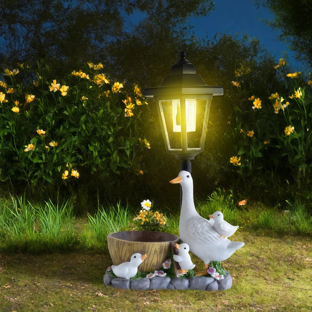 A glowing Solar Goose Lamp is accompanied by a goose and three goslings beside a wooden bowl, displayed on grass with yellow flowers and lush greenery in the background—an enchanting addition to your cottage garden décor.
