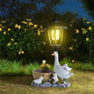 A glowing Solar Goose Lamp is accompanied by a goose and three goslings beside a wooden bowl, displayed on grass with yellow flowers and lush greenery in the background—an enchanting addition to your cottage garden décor.