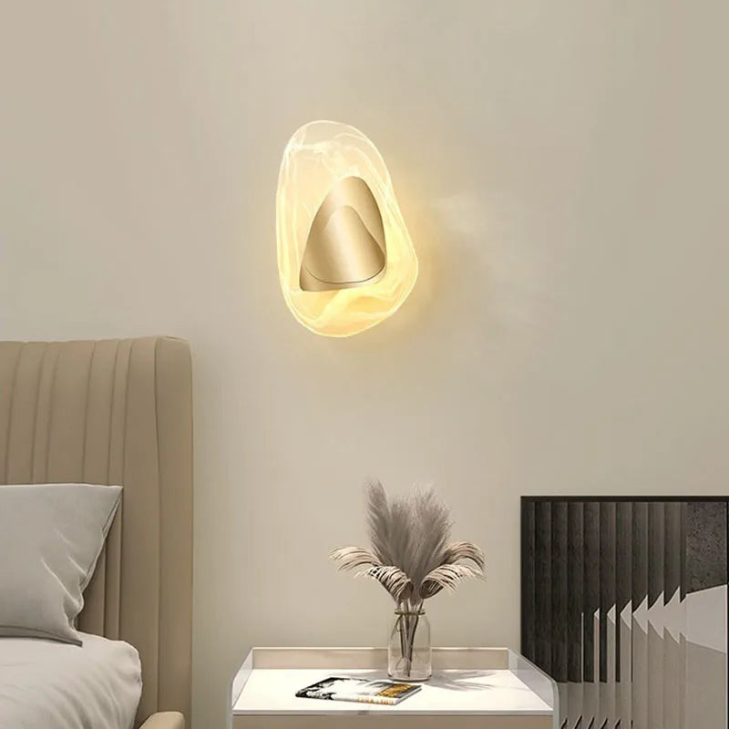 The Luxury Crystal Wall Lamp emits a warm glow above a nightstand adorned with a vase of dried plants, a magazine, and a decorative panel, positioned beside a beige upholstered bed.