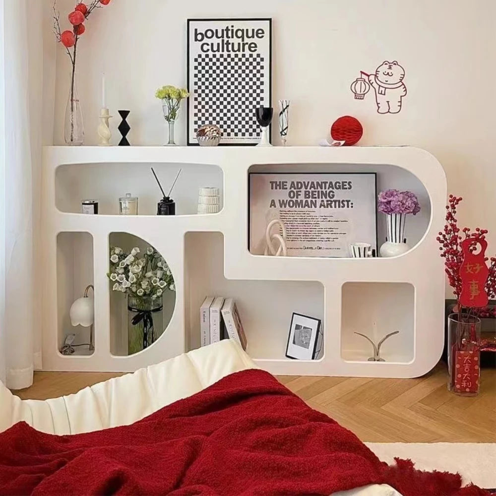 The Minimalist Cream Wind Sculptural Shelf, featuring geometric cutouts, stylishly displays books and decor. Art prints and vases on top add French cream wind charm. In front, a bed with red and white bedding completes the modern look.