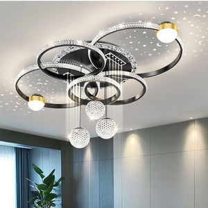 The Elegant Multi-Ring Ceiling Light features circular LED rings and crystal spheres that project star patterns onto the ceiling, enhancing a contemporary room with gray walls and a tall green plant.