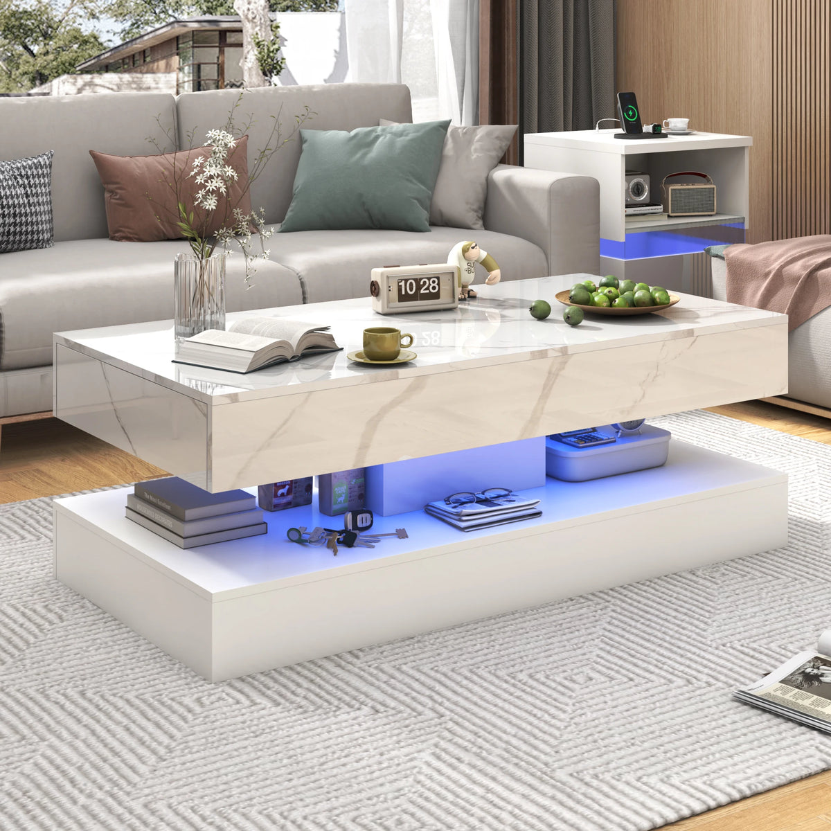 Modern living room showcasing the Premium Coffee Table with white marble finish, blue LED lighting, hidden storage, books, clock, plant, cup, green apples—paired with a gray sofa and textured rug.