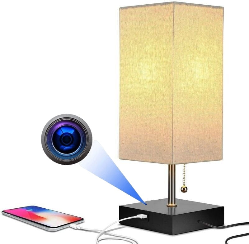The Desk Lamp Charging SpyCam, inspired by James Bond, is a rectangular table lamp with a light shade, USB charging port, and a subtle camera lens icon. An iPhone connects to its base using a charging cable.