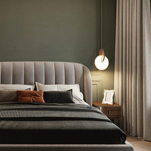 A bedroom featuring a gray upholstered headboard, pillows, and a plaid blanket exudes sophistication. Completing the scene is a modernist Dual Disc Pendant Light and a side table adorned with a frame and curtains.