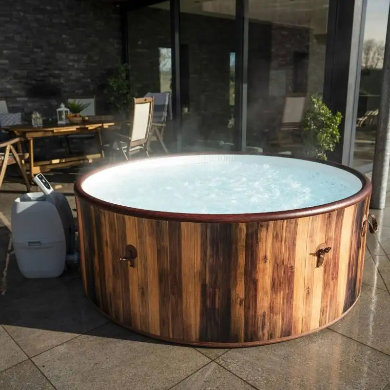 A round Outdoor Spa Hot Tub with bubbling water sits on a tiled patio near outdoor furniture and plants, positioned outside a modern house with large glass doors. This inviting Outdoor Spa Hot Tub creates the perfect backyard oasis, seamlessly blending relaxation and style for an exceptional outdoor spa experience.