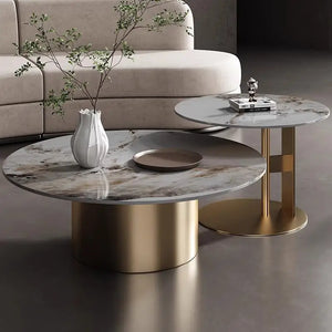 A beige sofa is fronted by two Premium Round Coffee Tables, one featuring a gold base and sintered stone top. One table holds a white vase with branches; the other displays a book and a small decorative item.