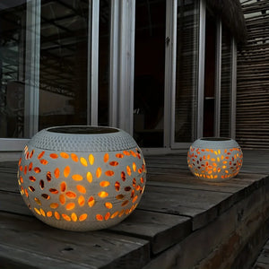 Two Solar Orb Lamps, featuring round white designs with leaf-shaped cutouts, sit on a wooden deck glowing warmly in front of glass windows and bamboo siding.