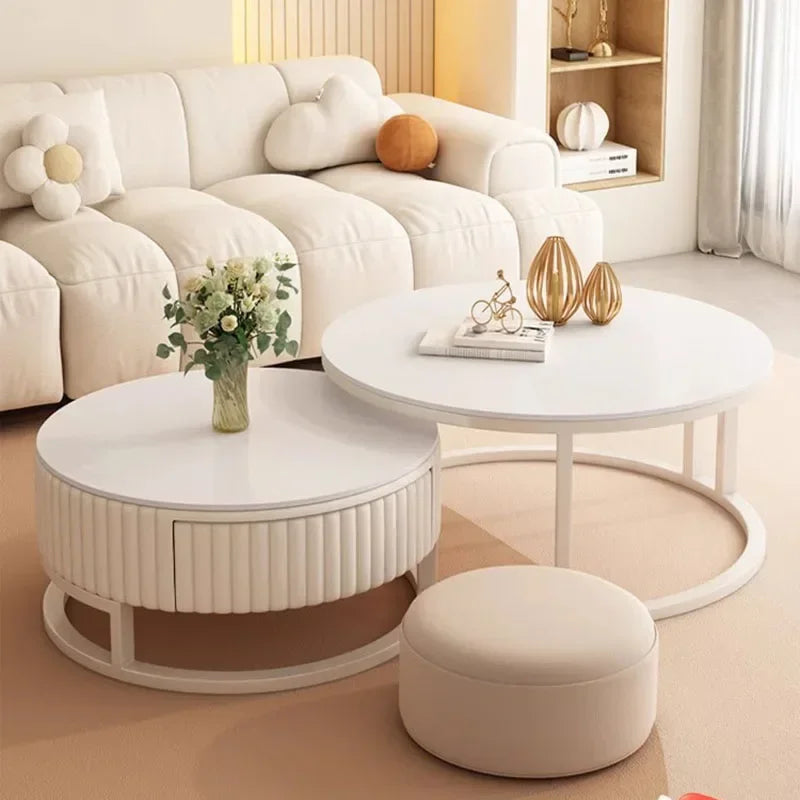 Modern living room with a white sectional sofa, a Luxury Round Coffee Table Set—one table with decor and books, another with a vase of flowers—and a round beige ottoman on a beige rug.