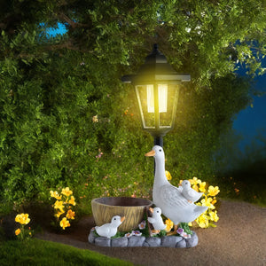 The Solar Goose Lamp illuminates a charming scene featuring a decorative goose with three goslings next to a small basin, all nestled among vibrant yellow flowers and lush greenery—an enchanting piece of garden decor ideal for any cottage garden.