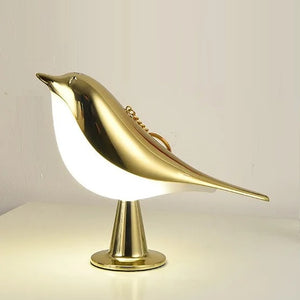 The Bird Table Lamp features a sleek, modern design with a gold finish and is perched on a round base, boasting an intuitive touch sensor for easy control.