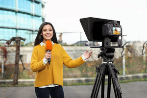 A reporter in a yellow sweater holds a microphone while standing outdoors, equipped with a camera setup on a tripod and using the Universal Portable Teleprompter for seamless content creation.