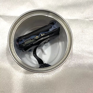 A damaged electronic component is placed inside a metal can on a shiny white surface, resembling the Fake Decorative Display SpyCam.