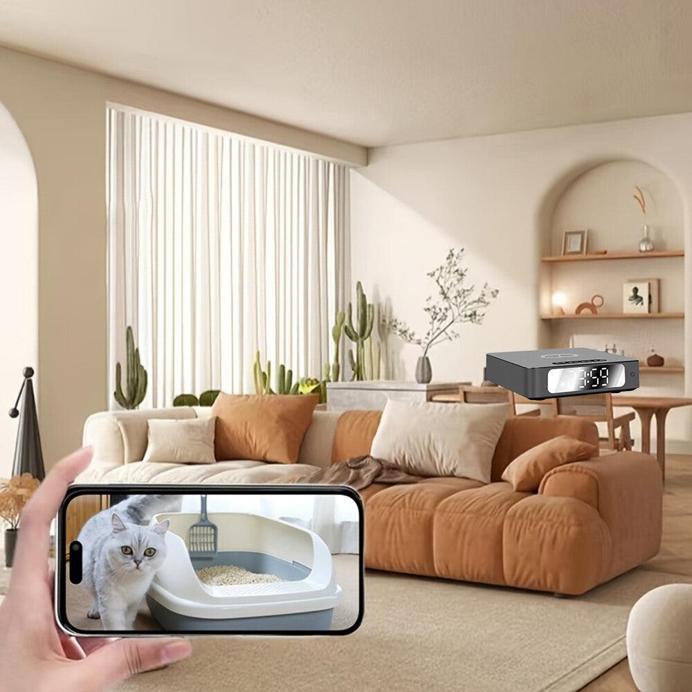 A hand holds a smartphone showing live video of a cat in a litter box, captured by the Wireless Charger Clock SpyCam with motion detection, set in a modern living room with a digital clock visible in the background.
