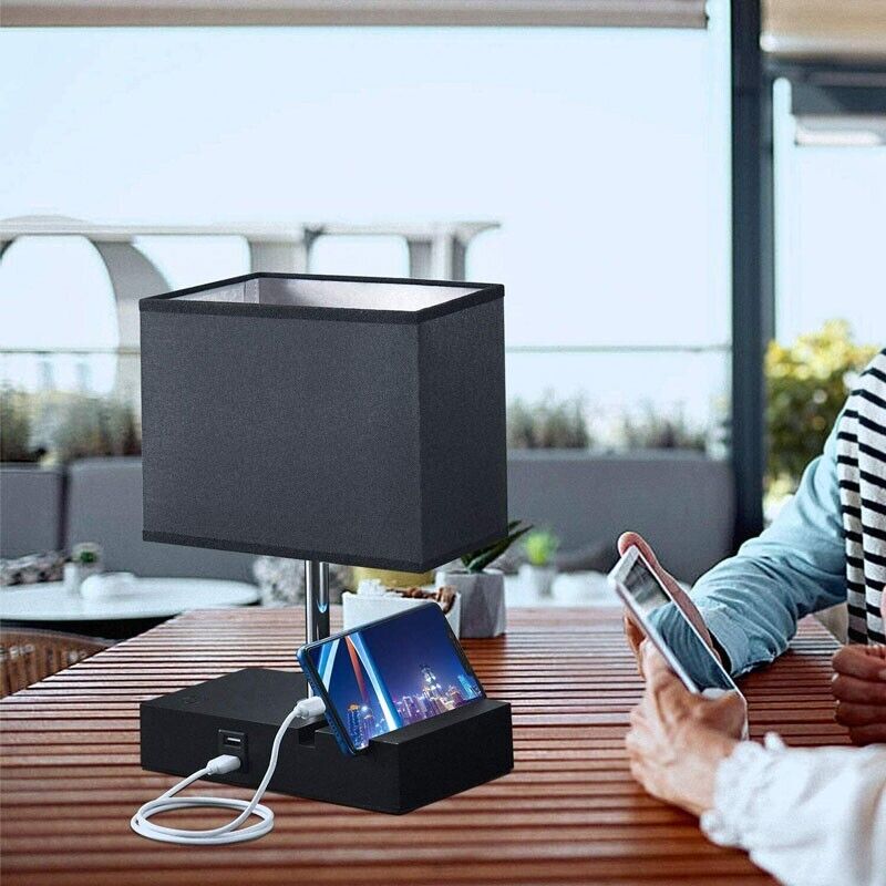 The Desk Lamp Charger SpyCam, with a rectangular shade and built-in USB charging port connected to a smartphone, sits elegantly on a wooden table. Nearby, someone uses another device while the lamp discreetly houses an HD 1080P SpyCam.
