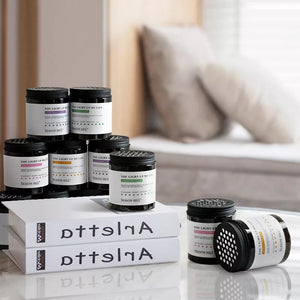 A stack of scented candles and books titled "Arietta" are displayed on a table, discreetly accompanied by the Cylindrical Display SpyCam that boasts 1080p HD resolution. The background includes a blurred bed and pillows.