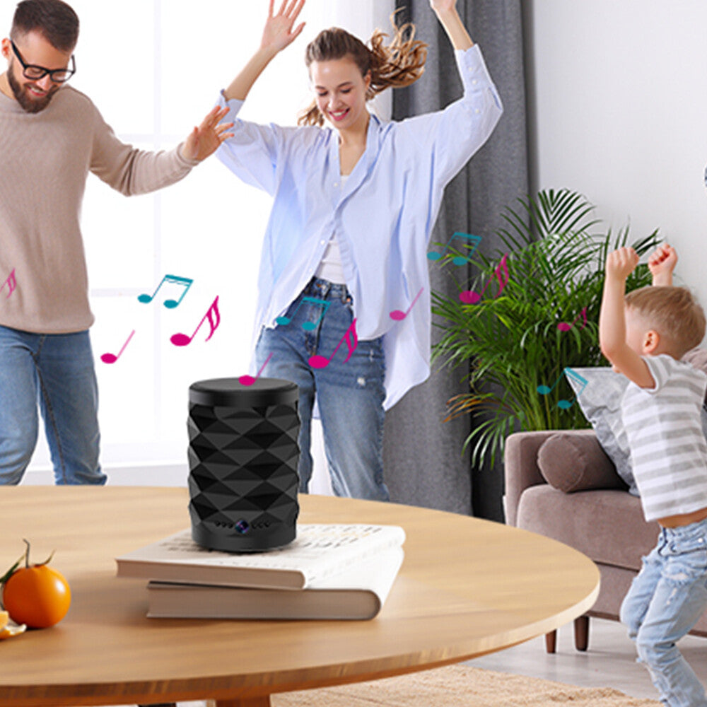 A man, woman, and child dance in a living room with music notes near a black Bluetooth Speaker SpyCam placed on a round wooden table.