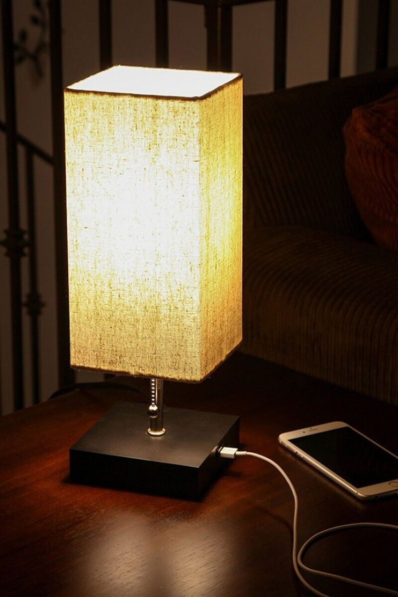 The Desk Lamp Charging SpyCam, inspired by James Bond, features a beige fabric shade that lights up the wooden surface where a smartphone connected to its charging cable rests nearby.