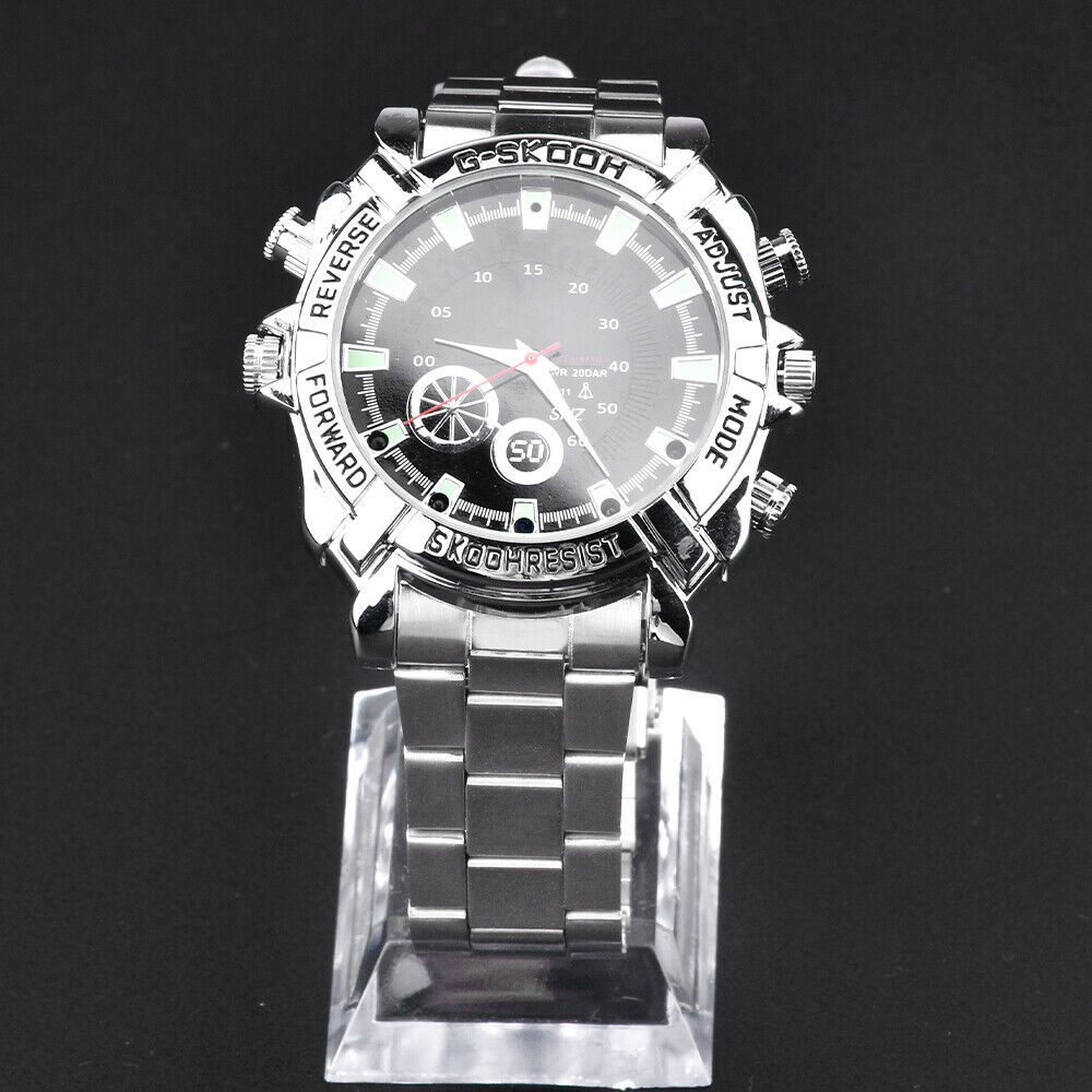 The Watch SpyCam is a silver metal wristwatch with a round black dial and buttons labeled "reverse," "forward," "adjust," and "mode." It discreetly records 1080P video and audio, shown on a clear stand against a black background.