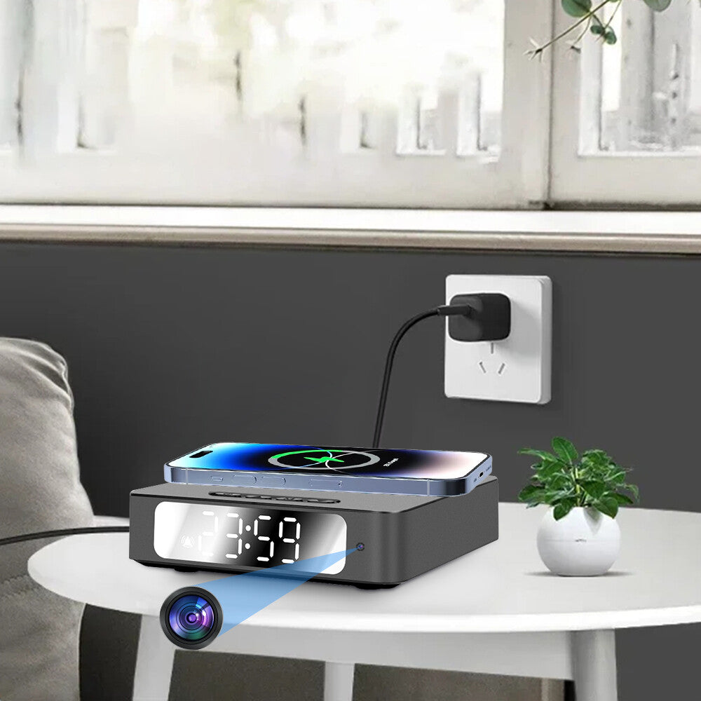 The Wireless Charger Clock SpyCam sits on a round table beside a potted plant and wall outlet, wirelessly charging a phone on top. A hidden spy camera lens is visible on the digital clock’s face.
