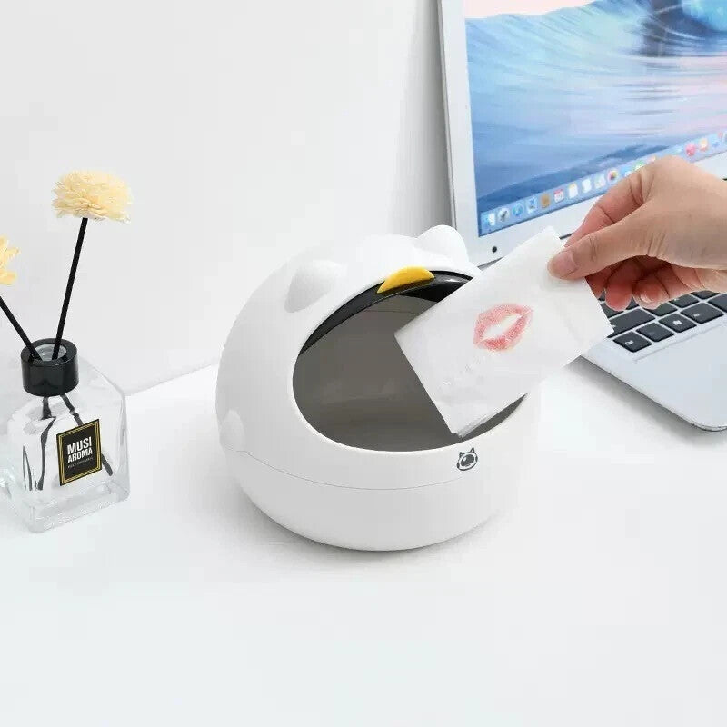 A hand puts a lipstick-marked tissue into the Cute Display SpyCam, a white kitty-shaped security camera trash bin on a desk beside a laptop and a small flower vase.