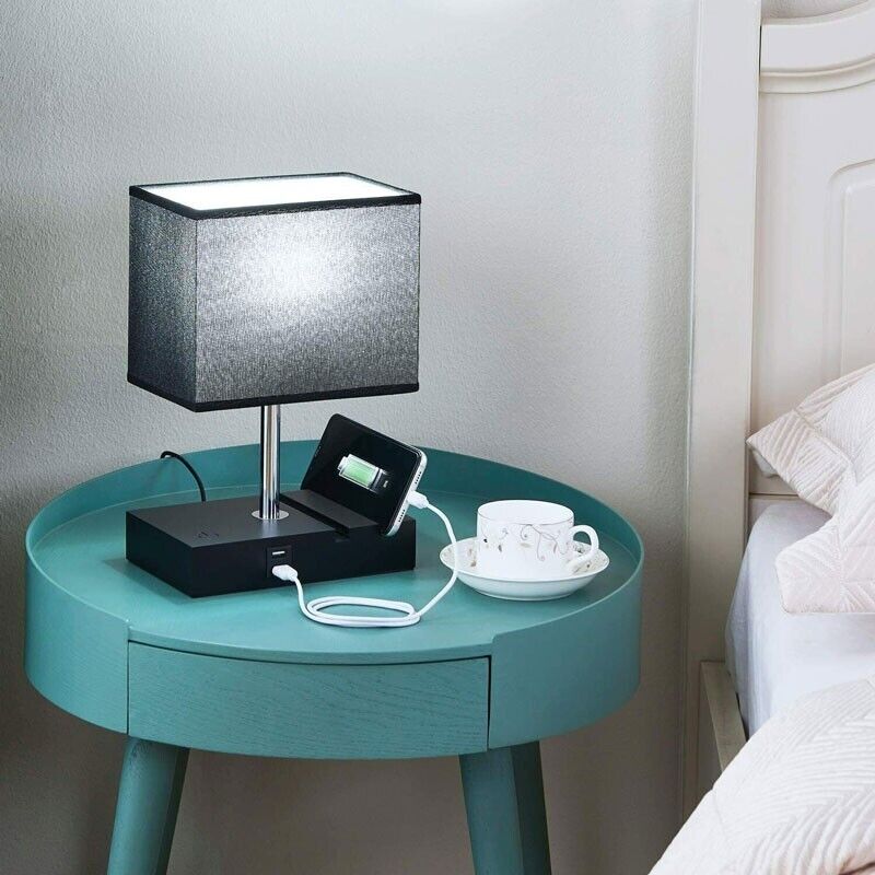 A Desk Lamp Charger SpyCam subtly oversees a bedside table, featuring a grey lamp, a smartphone charging dock, and a white cup and saucer for discreet surveillance in HD 1080P.